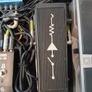 Černý efekt pedál s žebrovaným povrchem a bílým grafickým symbolem schématu. Zařízení je zapojeno do pedalboardu pomocí kabelů, z nichž jeden má žlut…
