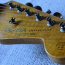 Detail hlavy elektrické kytary z přírodního dřeva. Na povrchu je černé logo Fender a nápis STRATOCASTER, pod ním text WITH SYNCHRONIZED TREMOLO PAT.…