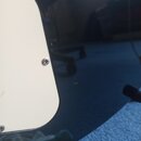 Detailní pohled na část těla elektrické kytary v černé barvě s lesklým povrchem. Na těle je přišroubován krémově bílý plastový plech (pickguard). Na…