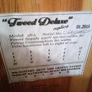 Fotografie detailu bílého papírového štítku s černým textem, který je nalepený na dřevěném povrchu. Štítek obsahuje nápisy „Tweed Deluxe“ replica, da…