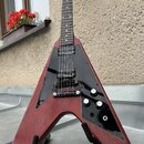 Elektrická kytara s Flying V tvarem těla v červeném dřevěném provedení a černým pickguardem. Na hmatníku jsou dva humbuckery, tři ovládací knofíky a…