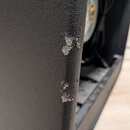 Detailní záběr na roh černého reproduktorového boxu s texturovaným povrchem. Na hraně je vidět výrazné opotřebení v podobě odřenin a odloupaného povr…