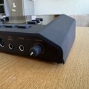 Černý elektronický hudební přístroj s ovládacími tlačítky na horní straně. Na zadním panelu jsou viditelné konektory a nápisy OUT, MIDI, IN, FOOT SWI…