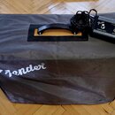 Šedý transportní kryt s bílým nápisem "fender". V horní části je otvor pro úchyt, skrze který je vidět oranžový povrch přístroje. Vedle krytu leží če…