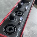 Červený audio rozhraní s černým horním panelem. Na panelu je nápis Scarlett 2i4, dva vstupy typu combo jack/XLR a ovládací prvky s popisky GAIN 1, GA…