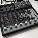 Šedý mixážní pult s černým rámem a ovládacími prvky v červené, modré a bílé barvě. Na horní hraně je nápis HEAINE 2. Zařízení obsahuje několik vstupn…