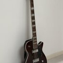 Tmavně hnědá elektrická kytara s polodupatým korpusem a bílým lemováním. Na hlavě hmatníku je viditelné logo GRETSCH. Kytara má dva snímače, stříbrný…
