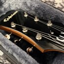 Detail hlavy černé kytary značky Epiphone, která leží v polstrovaném kufru. Na černé ploše je bílé logo Epiphone a bílý symbol připomínající stylizov…