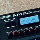 Detailní pohled na horní panel černého kytarového procesoru s modrým okrajem. Na zařízení je bílý nápis BOSS GT-1 GUITAR EFFECTS PROCESSOR. Podsvícen…