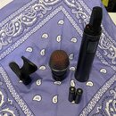 Na fialové látce s bílým vzorem je položen dynamický mikrofon s červenou mřížkou a černým tělem, na kterém je nápis V7 MG2. Vedle něj je černý bezdrá…