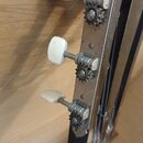 Detail hlavy strunového nástroje s černým povrchem a třemi ladicími mechanismy. Mechaniky mají kovové ozubené převody a bílé plastové knoflíky. Kovov…