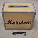 Hnědý kartonový box s rukojetí a nápisem Marshall AMPLIFICATION. Box je po obvodu polepen šedou lepicí páskou. Před krabicí leží černý napájecí kabel…