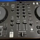 Černý DJ kontrolér s dvěma jog-koly a středovým mixážním sekcí. Na zařízení jsou viditelné nápisy DDJ-FLX4, rekordbox a serato, v dolní části je logo…