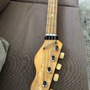 Detail hlavy basové kytary z přírodního dřeva. Na dřevě je svisle napsán text „natural bass“. Jsou vidět čtyři ladicí kolíky s bílými plastovými knof…