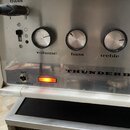 Detail přední strany zesilovače s kovovým panelem a bílým vinylovým potahem. Na panelu jsou tři ovládací knoflíky s popisky volume, bass a treble, př…