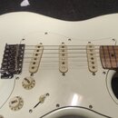 Bílá elektrická kytara typu Stratocaster s bílým pickguardem a hnedým hmatníkem. Nástroj má tři jednosmyčkové snímače v krémové barvě a chromovaný ko…