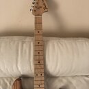 Elektrická kytara s přírodním dřevěným povrchem těla a hmatníkem. Na hlavě je černý nápis Fender STRATOCASTER. Nástroj má šest chromovaných ladiček a…