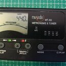 Černý digitální metronom a ladička s LCD displejem. Na přístroji je bílý nápis musedo MT-80 METRONOME & TUNER a označení MIC. Displej zobrazuje hodno…