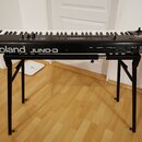 Černý digitální klavír umístěný na černém kovovém stojanu. Na přední straně je bílý nápis Roland JUNO-D a vpravo nahoře označení D-Synth keyboard. Ná…