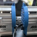 Dvě modré studiové lampy značky ARRI s označením 650W. Jsou vyrobeny z kovu s hliníkovým chladičem a mají černé úchyty. Na horní části je viditelný b…
