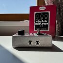 Stříbrný efekt pedál v kovovém šasi s dvěma vstupy/výstupy na zadní straně. Na horní části je vidět černý ovládací knoflík a červený panel s bílými n…