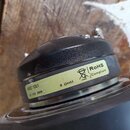 Fotografie zachycuje zadní část černého reproduktoru. Na spodním okraku je nalepený žlutý štítek s nápisy ASD 1001, 8 OHM a RoHS Compliant spolu s ik…