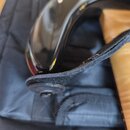 Detailní pohled na černý lakovaný trup nástroje s připojeným černým textilním popruhem. Popruh je upevněn pomocí kulatého knoflíku. Na povrchu popruh…