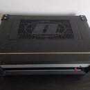 Černý zesilovač v transportním kufru s kovovými rohy a hranami. Na horní straně je ventilační mřížka s vyřezaným logem ve tvaru stylizovaného písmene…