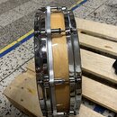 Fotografie zobrazuje snare drum s přírodním dřevěným povrchem korpusu a chromovaným kováním. Nástroj je umístěn na dřevěné paletě. Na viditelných čás…