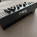 Černý syntetizátor s kovovými bočními panely v měděné barvě. Na pravém boku je bílý nápis moog. Horní panel obsahuje řadu černých otočných knoflíků a…
