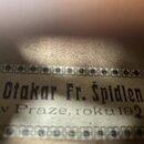 Detailní záběr na papírový štítek s černým textem a dekorativním rámečkem. Na štítku je nápis „Otakar Fr. Špidlen v Praze, roku 1925“. Papír vykazuje…