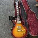 Elektrická kytara s trupem ve tvaru Les Paul v barevnosti sunburst, umístěná v otevřeném tvrdém kufru s červeným polstrováním. Na hlavě hmatníku je b…