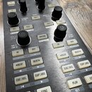 Detailní pohled na ovládací panel DJského kontroléru v černé barvě s kovovými prvky. Na panelu jsou umístěny černé otočné knoflíky a bílá tlačítka s…