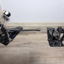 Černý dvojpedál k bicím s kovovým provedením a propojnou tyčí. Na pedálech nejsou viditelná žádná loga ani názvy modelů. Povrch je výrazně znečištěný…