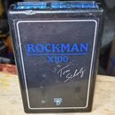 Černý kytarový procesor s bílým rámečkem. Na přední straně je modrý nápis ROCKMAN a pod ním označení X100. Vedle je bílý podpis Tom Scholz a ve spodn…