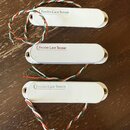 Tři bílé elektrické snímače s oválným tvarem a montážními šrouby po stranách. Každý snímač má na horní části nápis Fender-Lace Sensor. Ze snímačů ved…