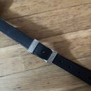 Černý kytarový popruh z kůže s kovovou přezkou. Na kovovém prvku je vyražen nápis FENDER. Povrch popruhu vykazuje mírné známky opotřebení a vrásnění.