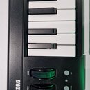 Černý MIDI kontrolér s bílými a černými klávesami. Na levé straně je bílý nápis KORG a dva ovládací knoflíky s popisky MOD a PITCH. Vedle nich jsou d…