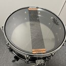 Fotografie znázorňuje snare drum s průhlednýmI skinem, na kterém je napnutý kovový snare-wire pás v měděné barvě. Na skinu je viditelné logo REMO s n…