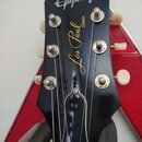 Černá hlava elektrické kytary s bílým logem Epiphone v horní části. Uprostřed je zlatý nápis Les Paul JUNIOR. Na hlavě jsou šest kovových ladiček s b…