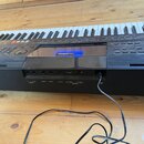 Černý digitální klavír s bílými a černými klávesami. Na horním panelu je modré logo YAMAHA. V zadní části jsou viditelné konektory s nápisy R OUTPUT,…
