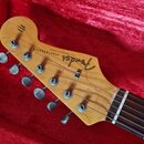 Fotografie zachycuje hlavu elektrické kytary z přírodního dřeva v červeném pouzdře. Na hlavě je černé logo Fender a nápis STRATOCASTER s menším texte…