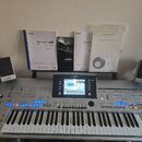 Stříbrný digitální klavír s nápisem YAMAHA a označením Tyros 4. Nástroj má v centru barevný displej, zahlreiche ovládací tlačítka a modře podsvícené…