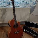 Akustická kytara s cutawayem v červenohnědé barvě, umístěná na černém stojanu. Na hlavě hmatníku je bílý nápis YAMAHA. Vedle nástroje leží otevřený č…