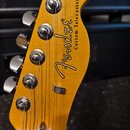 Detail hlavy elektrické kytary v žluté barvě s viditelnou strukturou dřeva. Na povrchu je černý nápis Fender® a pod ním menší text Custom Telecaster®…