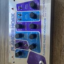 Kovový efekt pedál s nápisy FLANGER HOAX a electro-harmonix MADE IN USA. Zařízení má modré a fialové ovládací panely s několika černými otočnými knof…