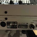 Záběr na zadní panel bílého syntetizátoru. Jsou zde vidět konektory s nápisy MIDI Out, MIDI Thru a B Outputs Right a Left. Na panelu jsou dvě nálepky…