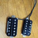 Dva černé humbuckery s bílými póly a připojeným černým kabelem. Na obou snímačích je bílý nápis Seymour Duncan. Snímače jsou umístěny na dřevěném pov…