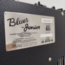 Detail stříbrného kovového štítku na černém texturovaném povrchu zesilovače. Na štítku je nápis Blues Junior a text A PRODUCT OF: FENDER MUSICAL INST…