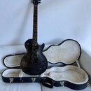 Černá elektrická kytara s tmavým dřevěným povrchem a v černé hardshell case s bílou vnitřní výplní. Na hlavě nástroje je zlatý nápis Gibson. Kytara m…